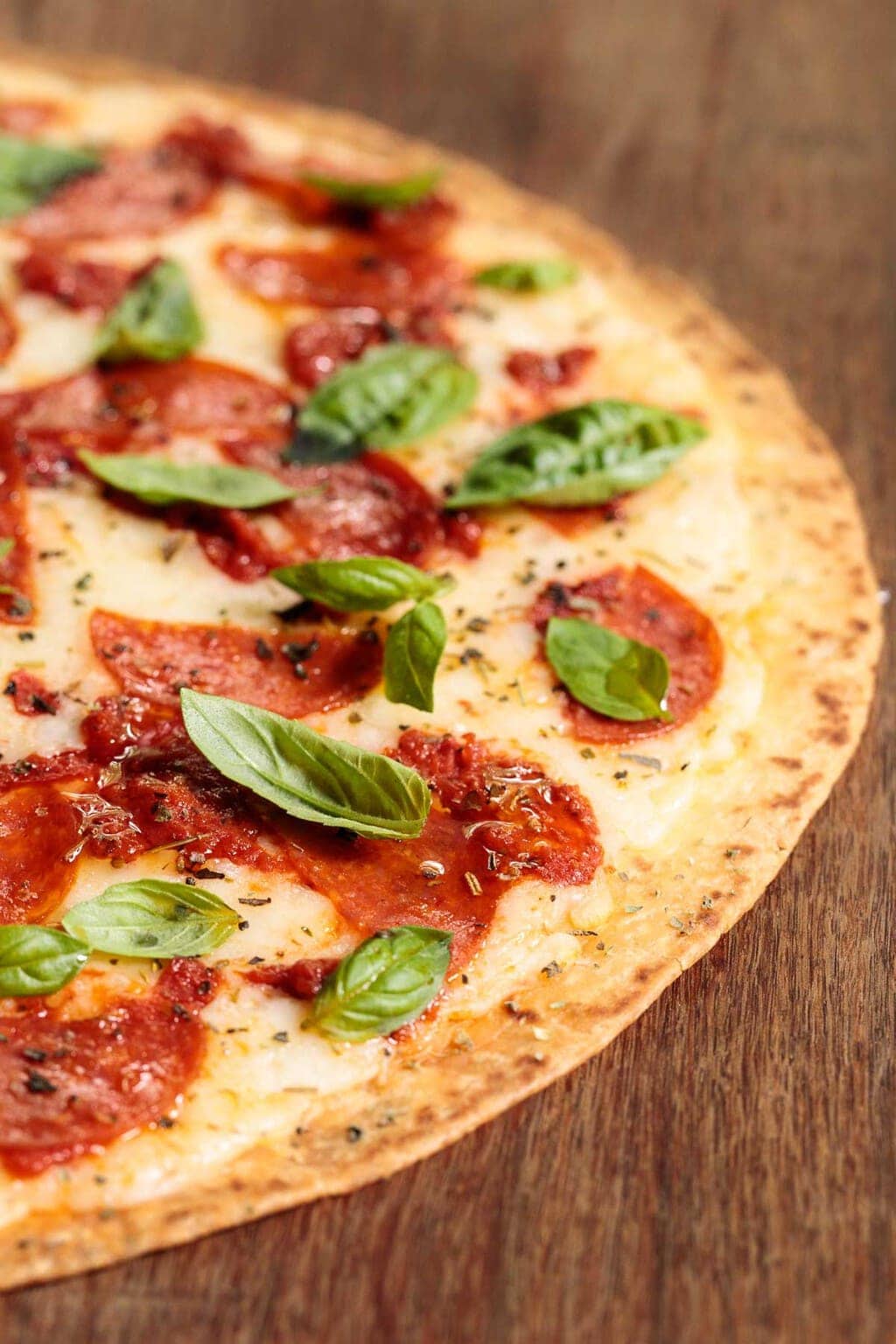 Closeup vertical photo of Easy Thin Crust Pizza with basil and pepperoni garnished with fresh basil leaves on a wood table.
