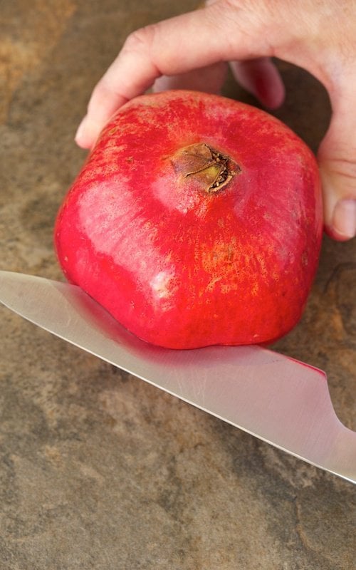 Vertical closeup photo of slicing a pomegranate in half demonstrating The Easy Way to Remove Pomegranate Seeds.