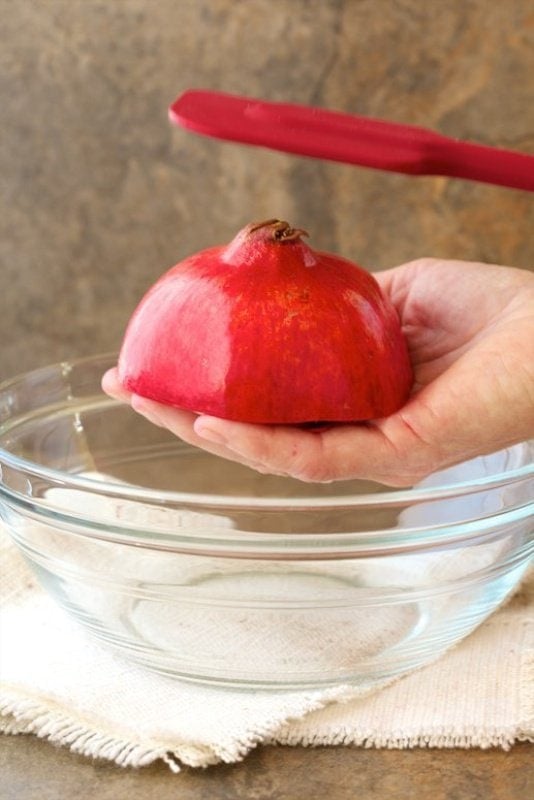 Vertical closeup photo of half of a pomegranate being struck with a spatula demonstrating The Easy Way to Remove Pomegranate Seeds.
