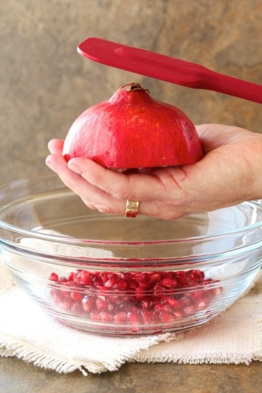 Vertical closeup photo of more of the arils falling into the glass bowl demonstrating The Easy Way to Remove Pomegranate Seeds.