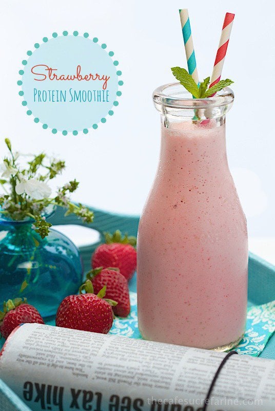 Photo of a glass carafe filled with a Strawberry Protein Smoothie. A morning newspaper, fresh strawberries and a flower in a vase are in the foreground. A graphic title is in the background.