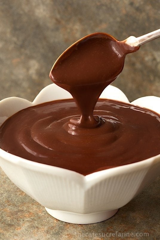Vertical photo of Best Ever Hot Fudge Sauce in a white scalloped bowl with a spoon pouring the sauce into the bowl.