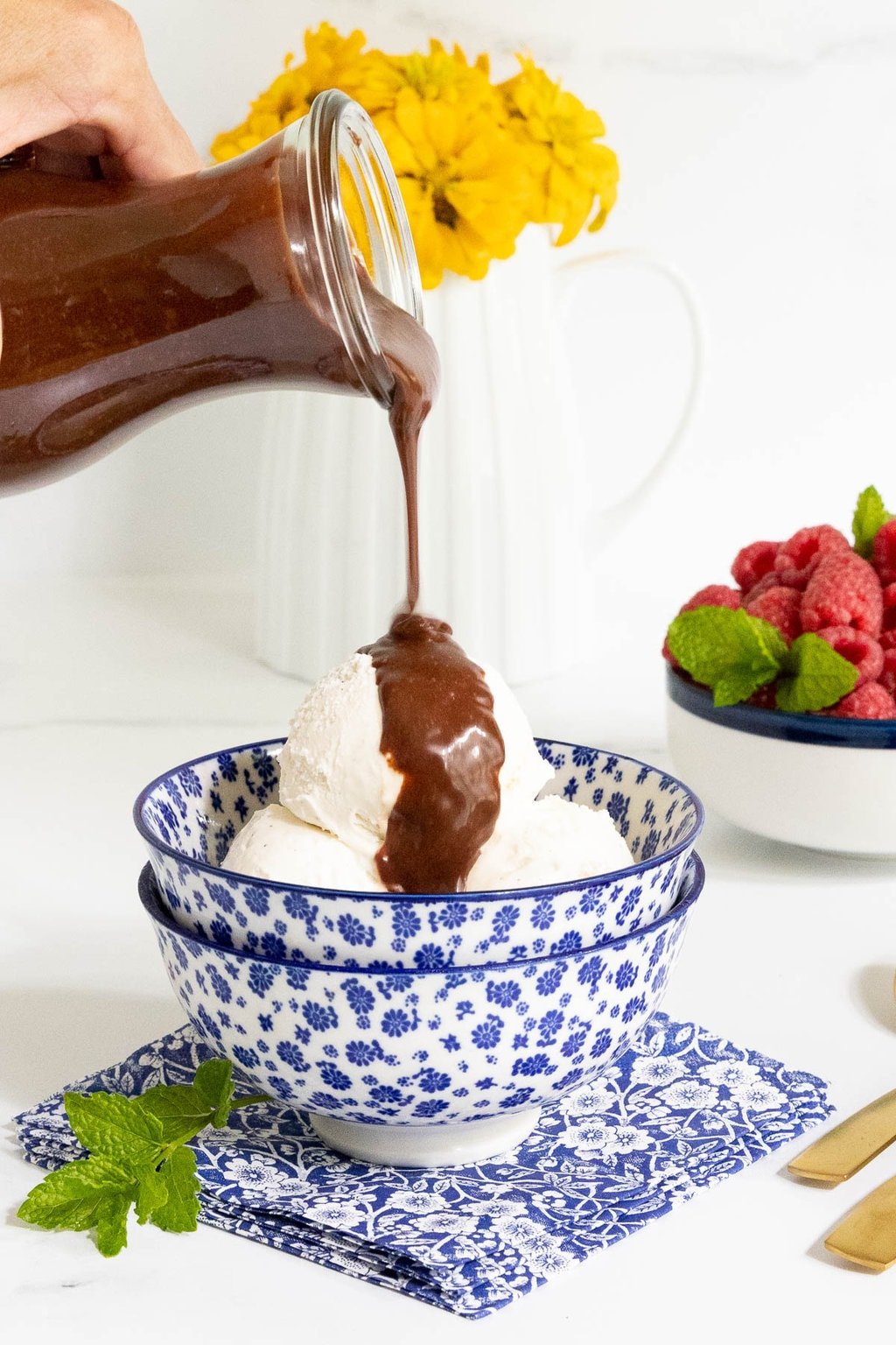 Vertical photo of a person pouring Ridiculously Easy Hot Fudge Sauce over a bowl of vanilla ice cream.