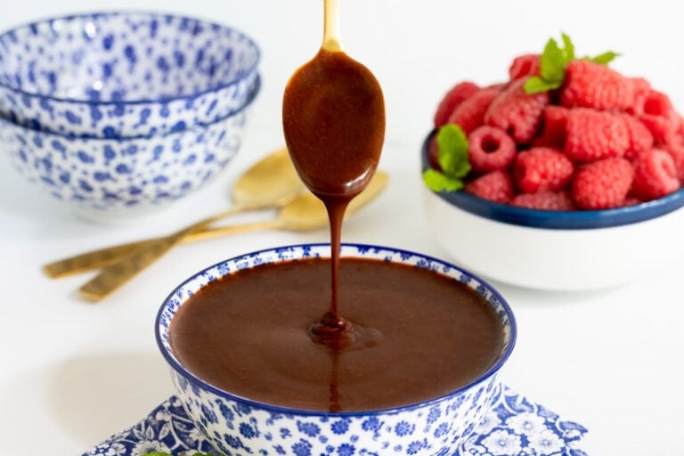 Horizontal photo of a bowl of Ridiculously Easy Hot Fudge Sauce with a spoon dripping the sauce back into a blue and white bowl.