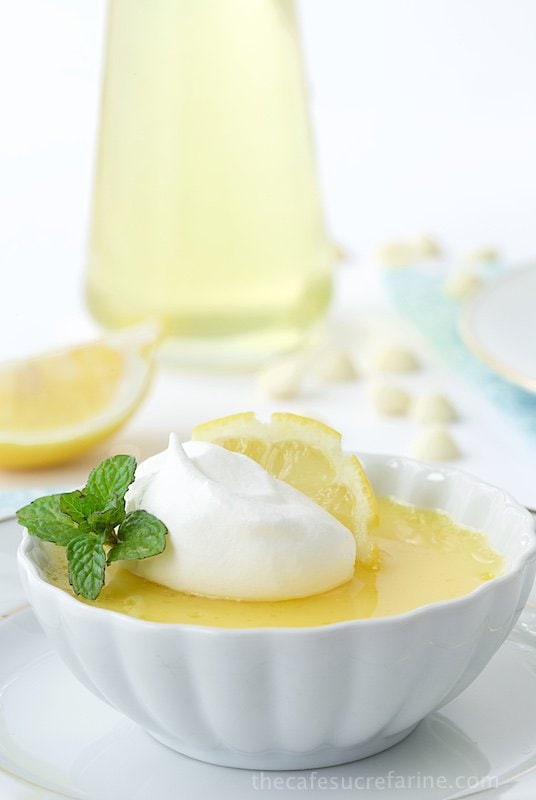 Vertical photo of a Lemon and White Chocolate Pots de Crème in a white ramekin with lemon slices, white chocolate chips and Limoncello sauce in the background.