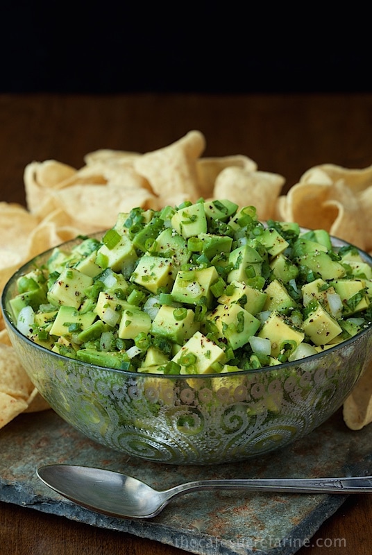 Photo of an etched glass serving bowl of Avocado Salsa on a slate surface with tortilla chips in the background.