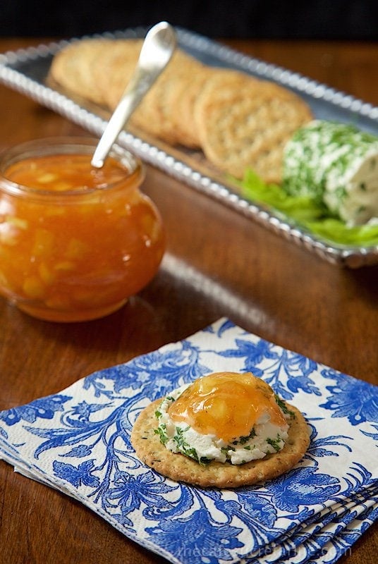 Photo of Pineapple Sriracha Freezer Jam on an appetizer cracker with cheese spread. A jar of the jam and an appetizer tray are in the background.