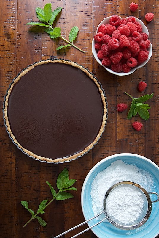 Overhead picture of French Chocolate Tart with Brown Butter Crust on a wooden table with raspberries and powdered sugar