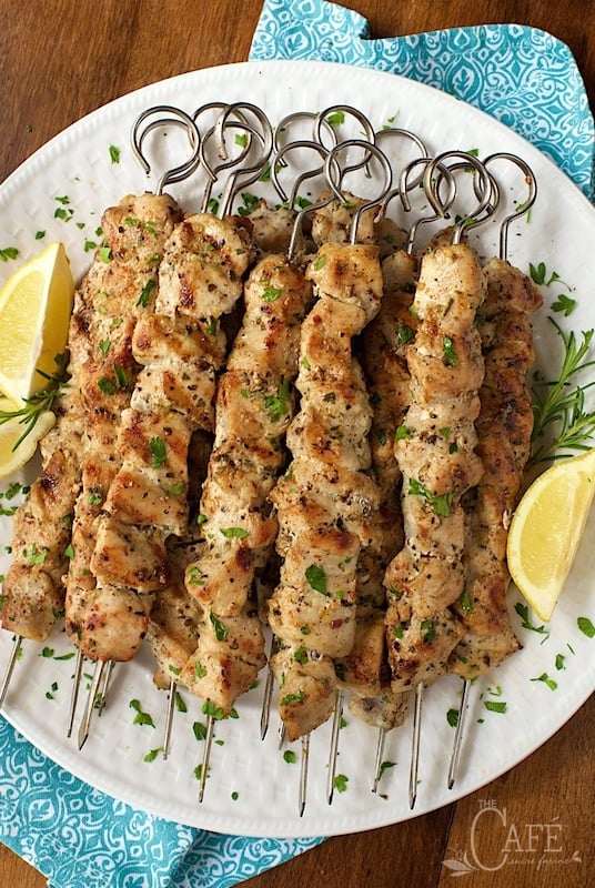 Overhead vertical photo of Lemon, Rosemary and Garlic Chicken Skewers on a white plate with lemons and herbs.