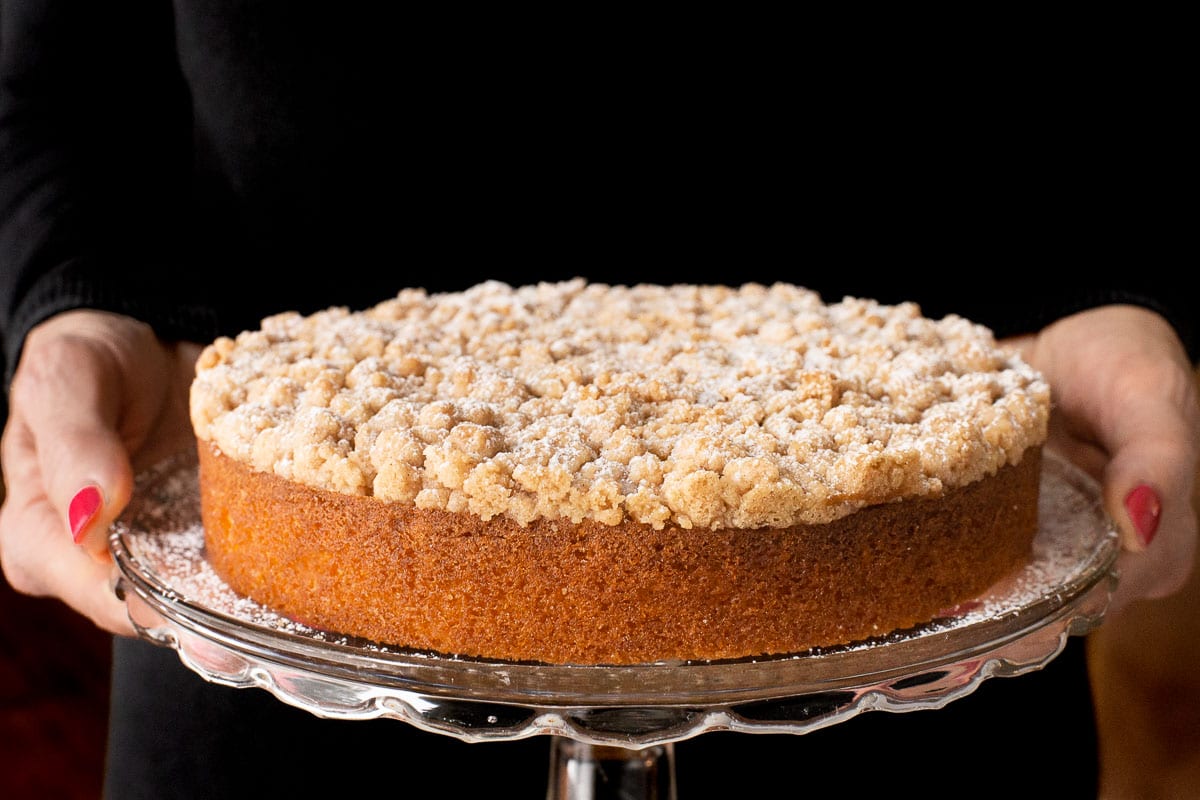 Horizontal closeup photo of a person holding a Easy Overnight Coffee Cake on a glass pedestal platter.