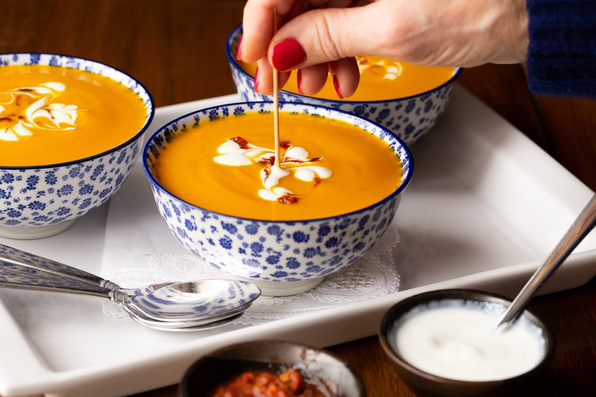 Horizontal closeup photo of Roasted Thai Carrot and Sweet Potato Soup in blu and white patterned serving bowls with a person decoratively stirring in Sriracha sauce.