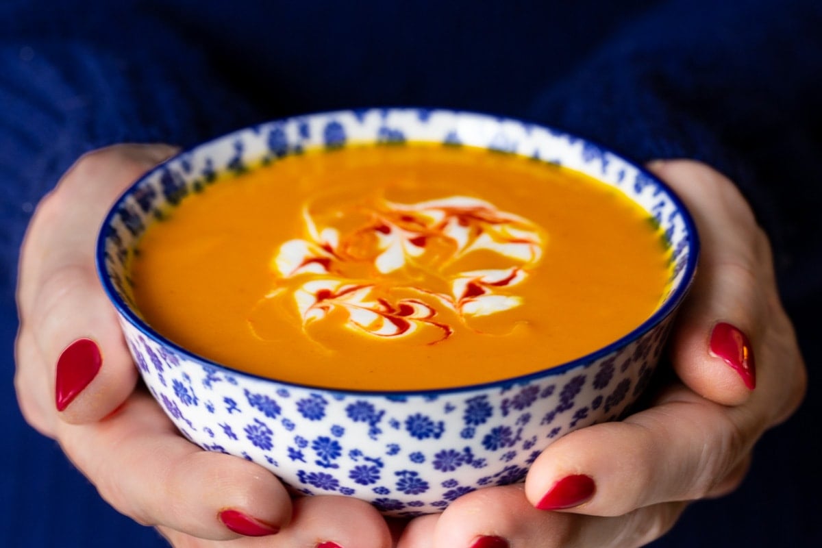 Horizontal closeup photo of a person holding a bowl of Thai Roasted Carrot and Sweet Potato Soup in a blue and white soup bowl.