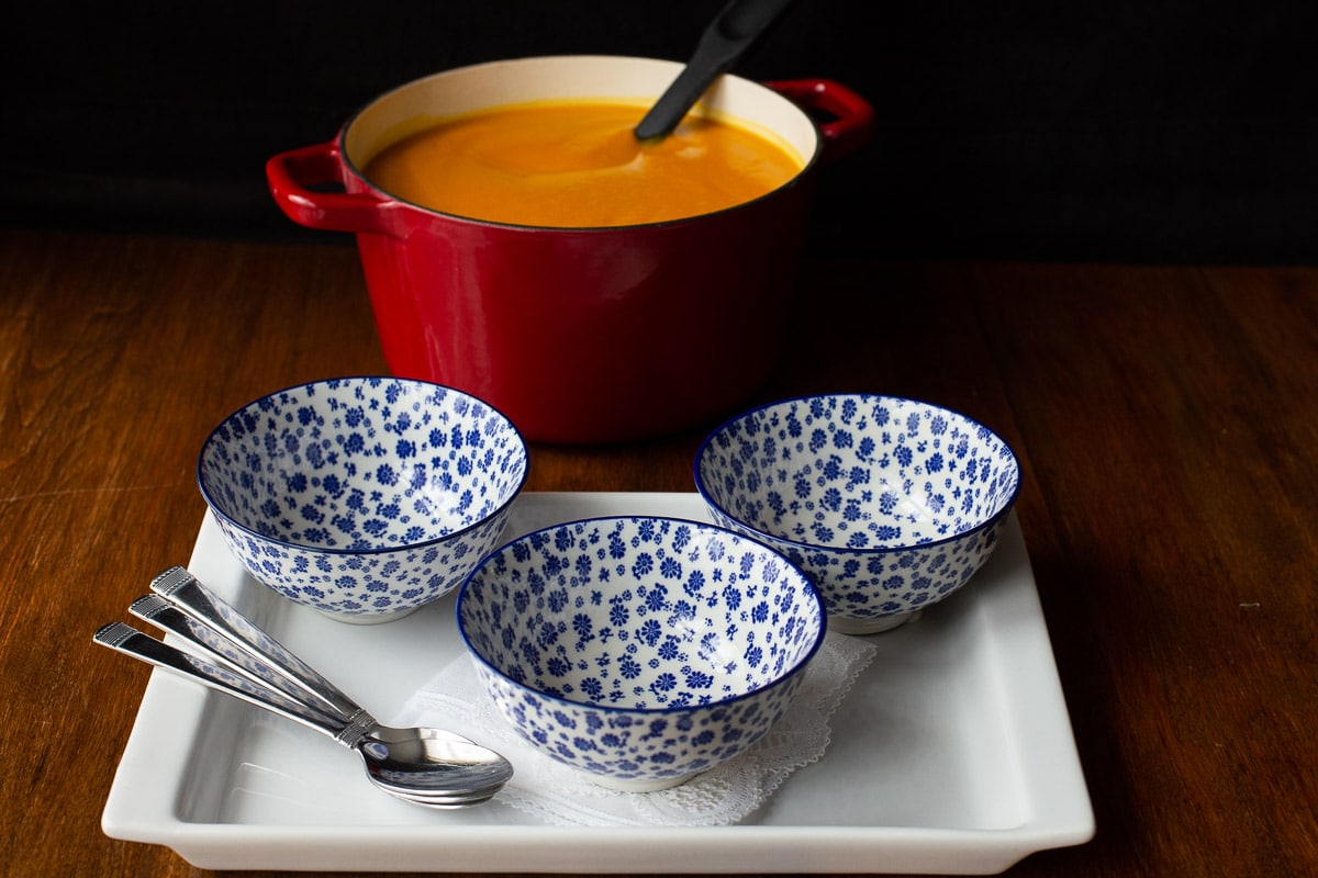 Horizontal photo of three empty blue and white patterned soup bowls and a red porcelain pot of Roasted Thai Carrot and Sweet Potato Soup.