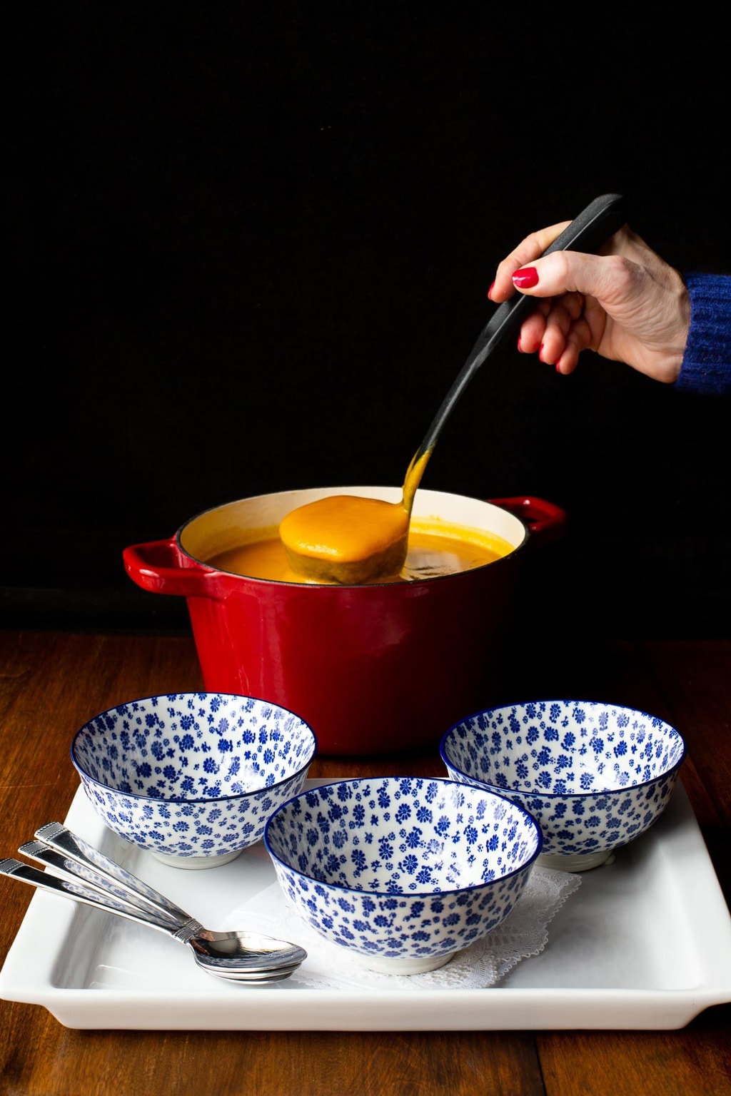 Vertical photo of a person ladling Roasted Thai Carrot and Sweet Potato Soup out of a red pot into smaller soup serving bowls.