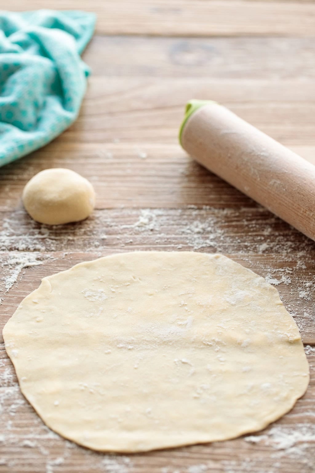 Rolled-out flour tortilla dough on a floured wooden surface with a dough ball, rolling pin, and kitchen towel in the background—step in making homemade tortillas from scratch.