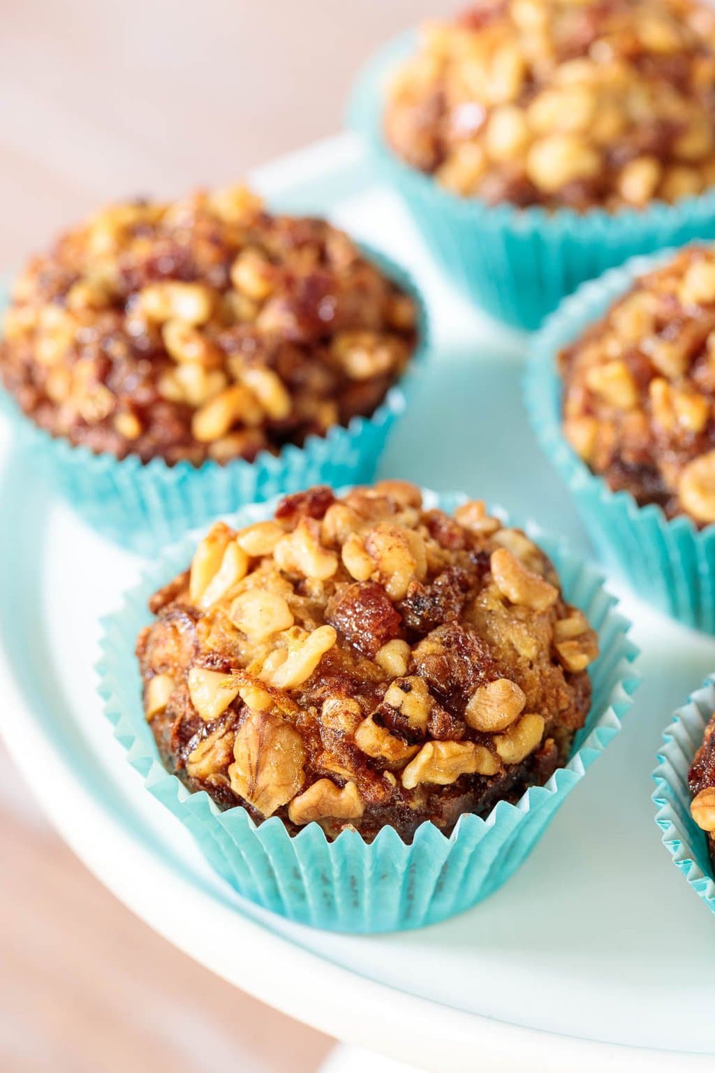 Vertical closeup photo of a batch of Candied Walnut Date Banana Muffins in turquoise cupcake liners on a white pedestal serving plate.