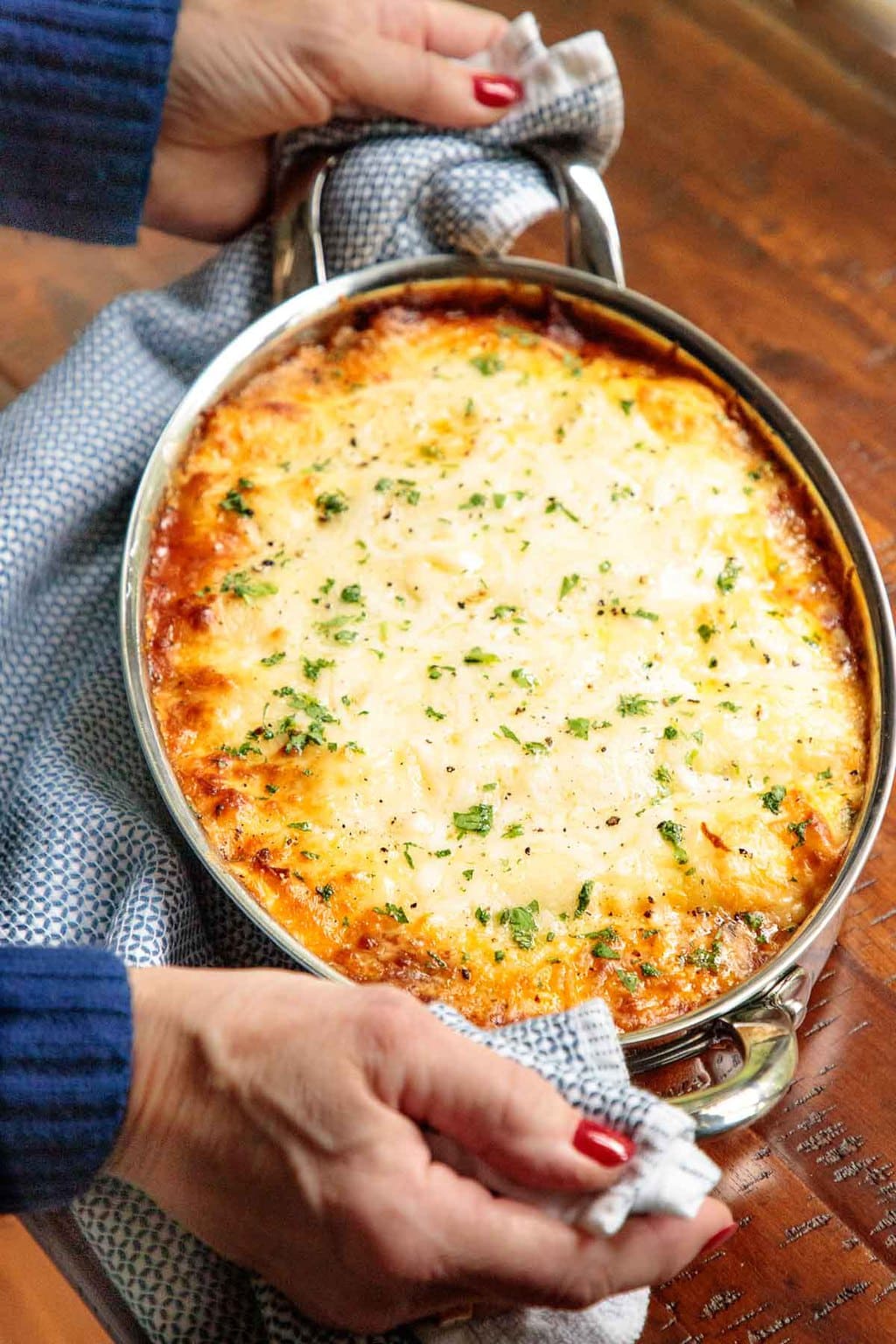 Vertical photo of a person holding a baking pan of Make-Ahead Homemade Manicotti above a wood table.