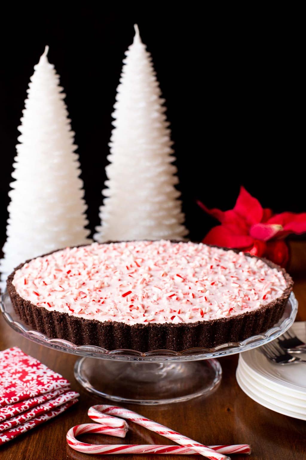 Vertical photo of a Peppermint Candy Cane Tart on a glass pedestal serving plate with wax Christmas tree candles in the background.