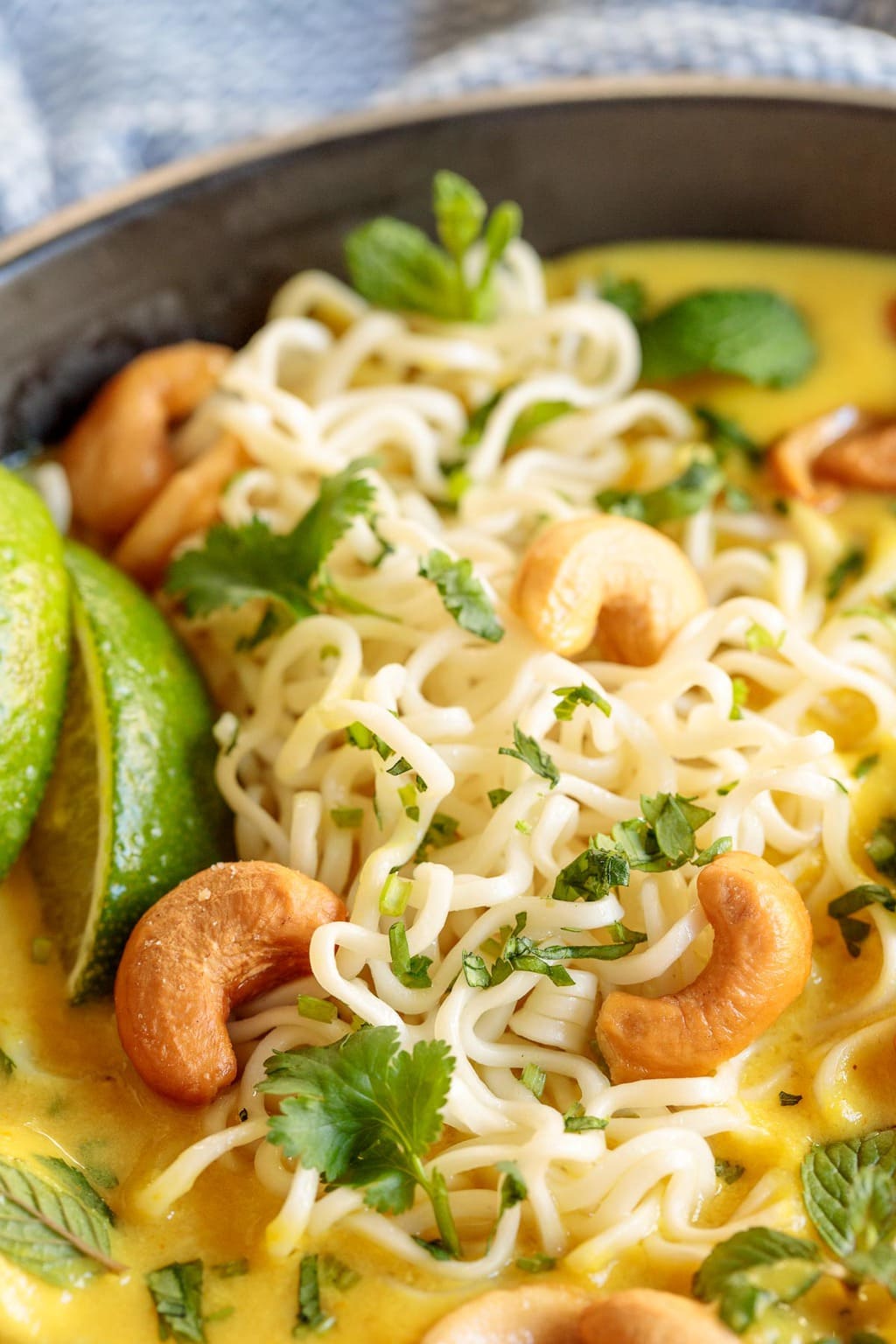 Vertical extreme closeup photo of a bowl of Coconut Curry Chicken Ramen garnished with lime wedges, cashews and fresh herbs.