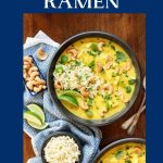 Vertical overhead photo of two bowls of Coconut Curry Chicken Ramen on a wood table surrounded by cashews, a small bowl of ramen noodles and lime wedges.