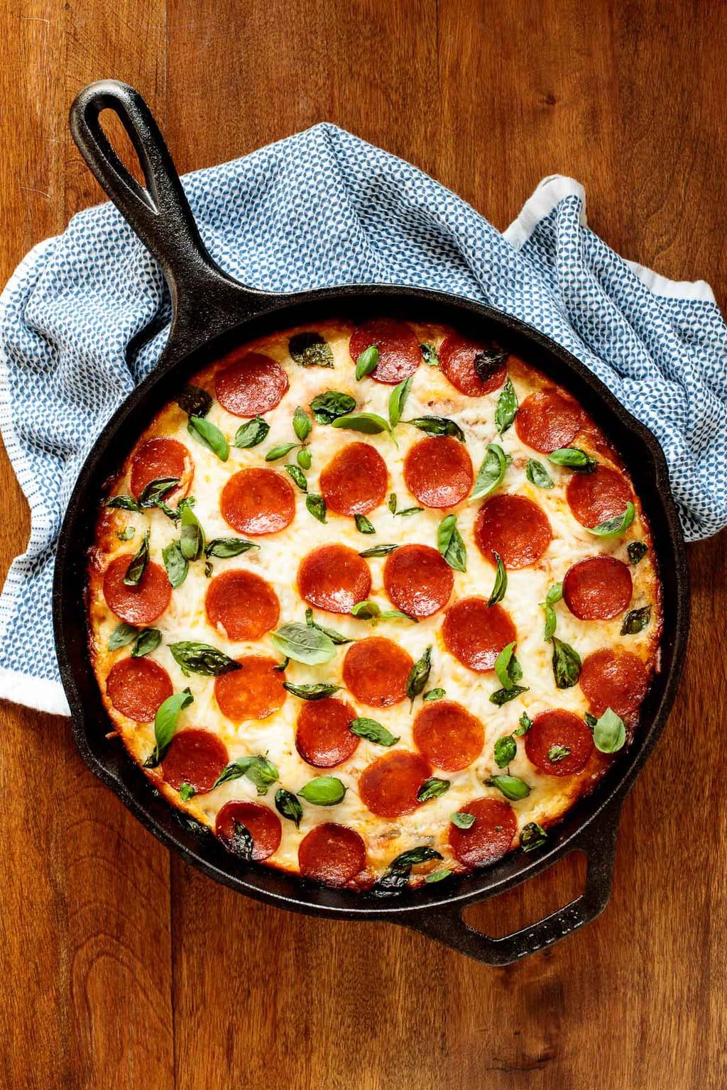 Vertical overhead photo of a cast iron skillet with a Deep Dish Pepperoni Basil Pizza on a wood table.