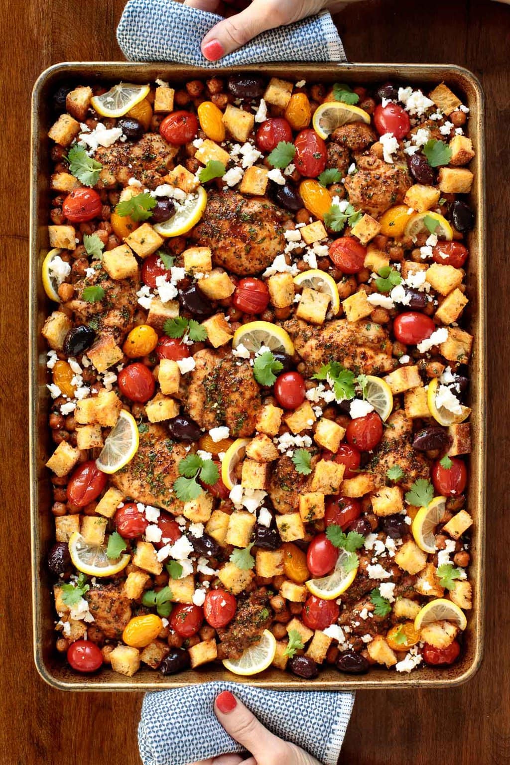 Vertical overhead photo of a person holding a finished pan of Mediterranean Sheet Pan Chicken .