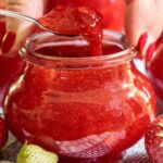 Spoonful of vibrant strawberry freezer jam being lifted from a glass jar, surrounded by fresh strawberries.