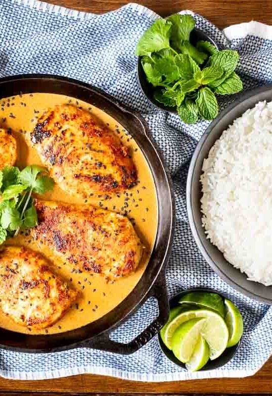Horizontal overhead photo of Coconut Braised Chicken Breasts in a cast iron skillet with bowls of jasmine rice, fresh herbs and lime wedges nearby.