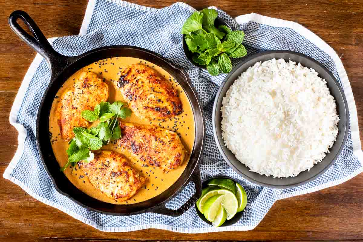 Horizontal overhead photo of Coconut Braised Chicken Breasts in a cast iron skillet with bowls of jasmine rice, fresh herbs and lime wedges nearby.