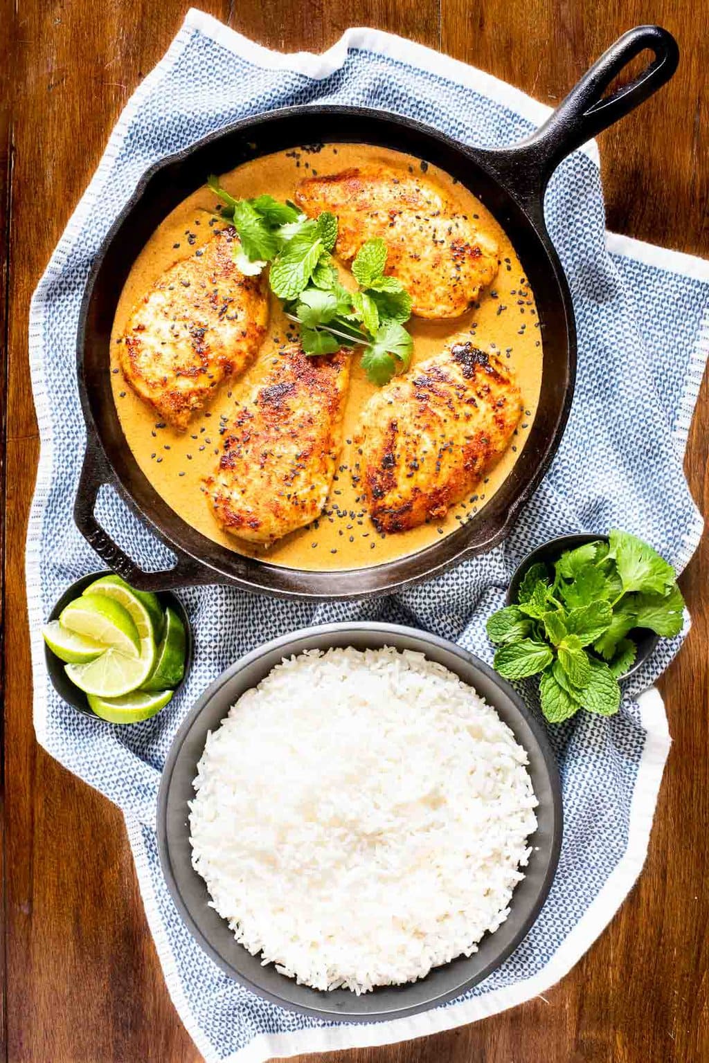 Vertical overhead photo of Coconut Braised Chicken Breasts in a cast iron skillet on a wood table next to a pan of jasmine rice.