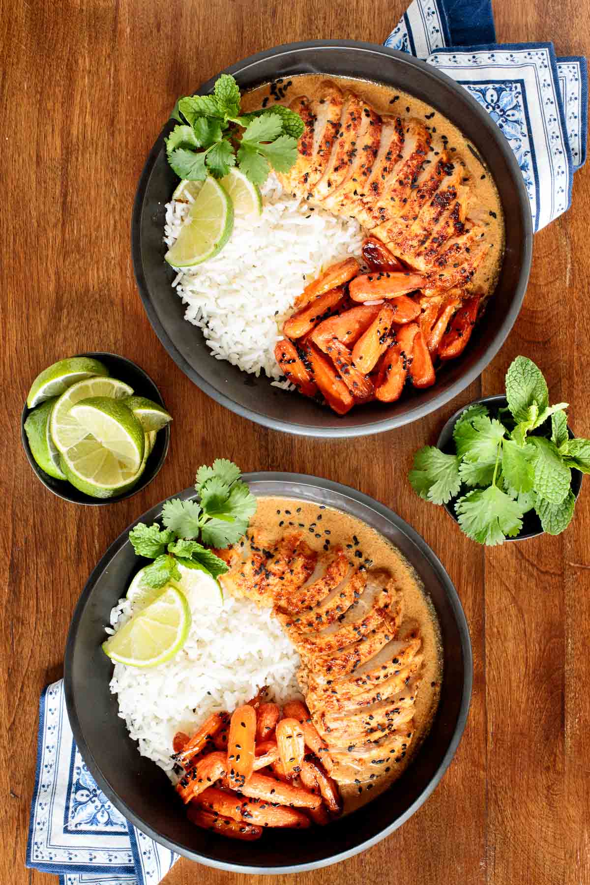 Vertical overhead photo of Coconut Braised Chicken Breasts in two black serving bowls with honey ginger charred carrots, jasmine rice, lime wedges and fresh herbs.