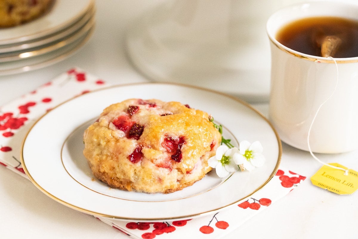 Horizontal photo of a Ridiculously Easy Fresh Strawberry Scone on a white and gold rimmed serving plate with a cup of tea.