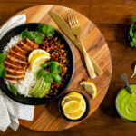 Overhead horizontal photo of Peruvian Chicken Rice Bowls surrounded by smaller bowls of lemon wedges, fresh basil leaves and Peruvian Green Sauce on a wood table.
