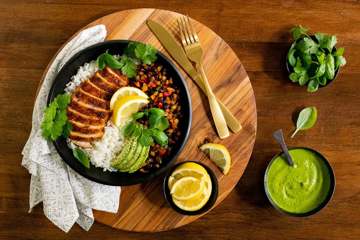 Horizontal overhead photo of Peruvian Chicken Rice Bowls on a wood table with fresh basil leaves, sliced lemon wedges and Peruvian Green Sauce.