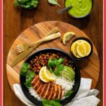 Vertical overhead photo of a Peruvian Chicken Rice Bowl on a round wood platter surrounded by smaller bowls of fresh basil leaves, Peruvian Green Sauce and lemon wedges with a text graphic at the bottom of the photo.