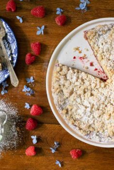 Overhead horizontal photo of a Easy Plum Raspberry Crumb Tart on a wood table surrounded by fresh raspberries and flowers.