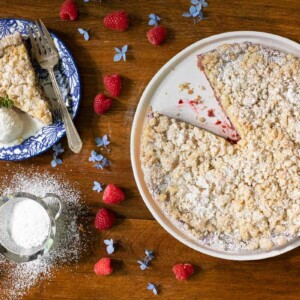 Overhead horizontal photo of a Easy Plum Raspberry Crumb Tart on a wood table surrounded by fresh raspberries and flowers.