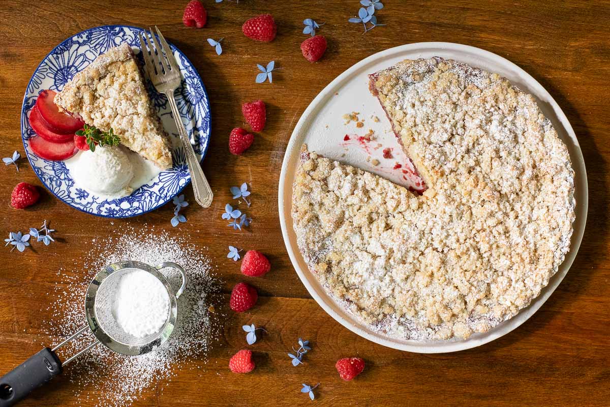 Overhead horizontal photo of a Easy Plum Raspberry Crumb Tart on a table with powdered sugar, fresh raspberries and flowers surrounding the tart.