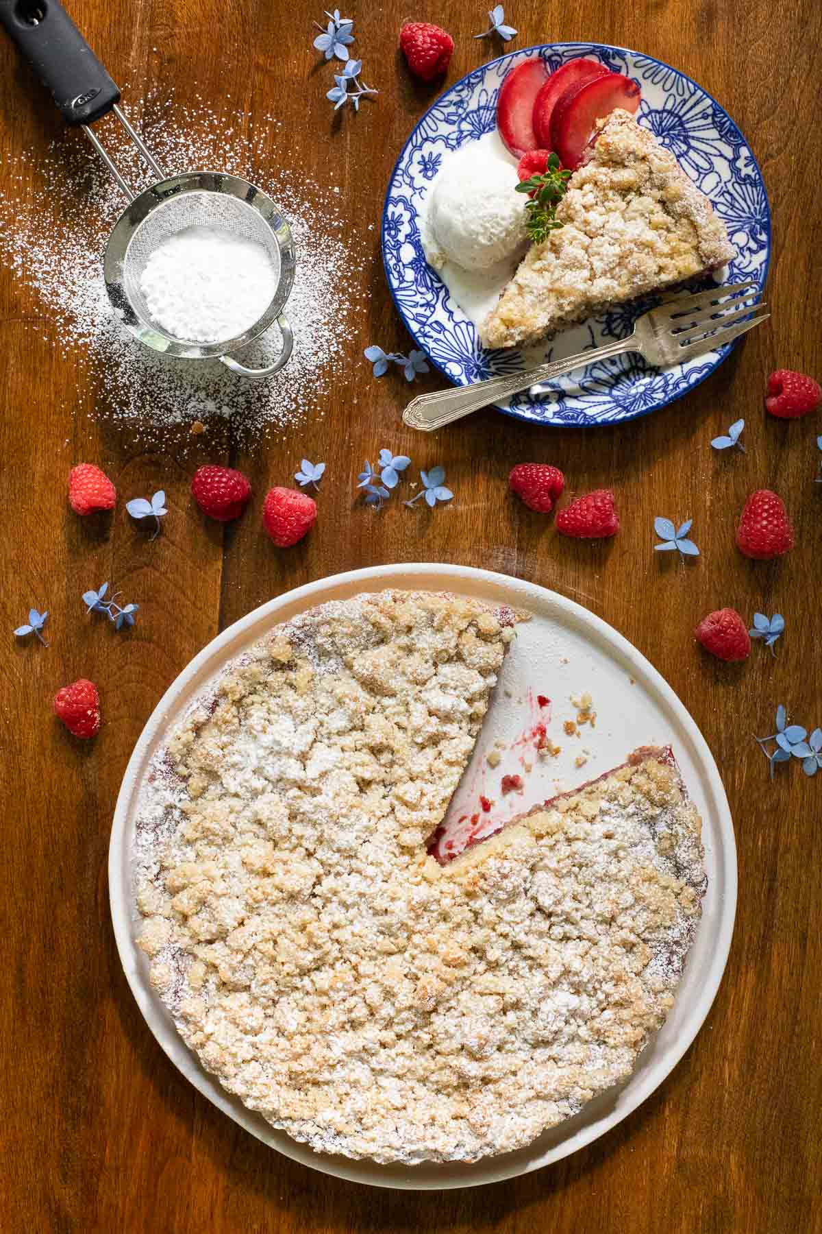 Vertical overhead photo of a Easy Plum Raspberry Crumb Tart surrounded by fresh raspberries, flowers and a powdered sugar sieve on a wood table.