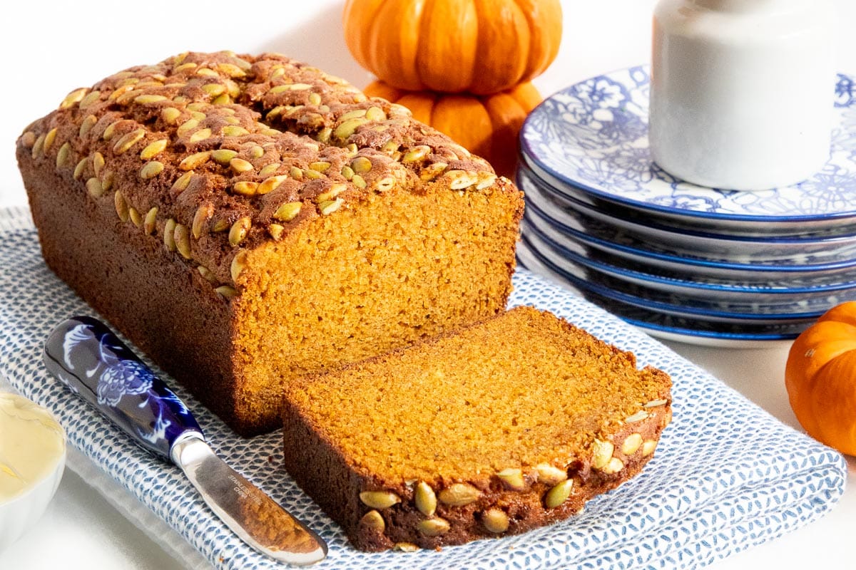 Horizontal closeup photo of a loaf of Pumpkin Bread on a blue and white cloth with mini pumpkins in the background.