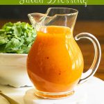 Vertical photo of a glass pitcher of Red Pepper Jelly Salad Dressing on a round marble surface with a baby arugula salad in the background.