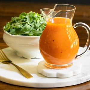 Horizontal closeup photo of a glass pitcher of Red Pepper Jelly Salad Dressing on a round marble surface with a lettuce salad in the background.