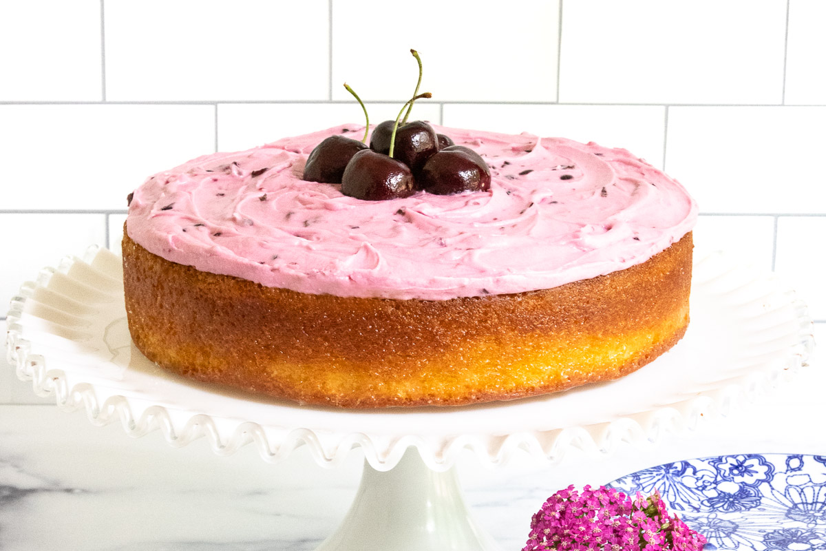 Horizontal photo of anEasy French Almond Cake with Fresh Cherry Buttercream on a white glass pedestal cake stand.