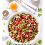 Vertical overhead photo of a Watermelon Arugula Salad with Pepper Jelly Dressing on a white marble surface with a bowl of Easy Candied Pecans.