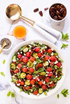 Vertical photo of photo of a Watermelon Arugula Salad with Pepper Jelly Dressing in a serving bowl on a white marble surface.