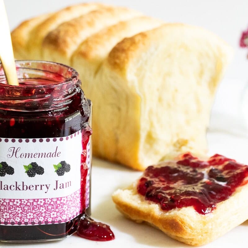 blackberry freezer jam spread on a piece of homemade toast with a custom labeled jar sitting beside the loaf