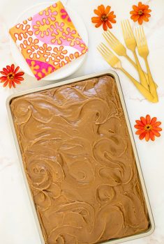 Vertical photo of a Ridiculously Easy Caramel Buttermilk Sheet Cake on a white marble surface. The photo has an orange border and the cake is surrounded by orange zinnias.
