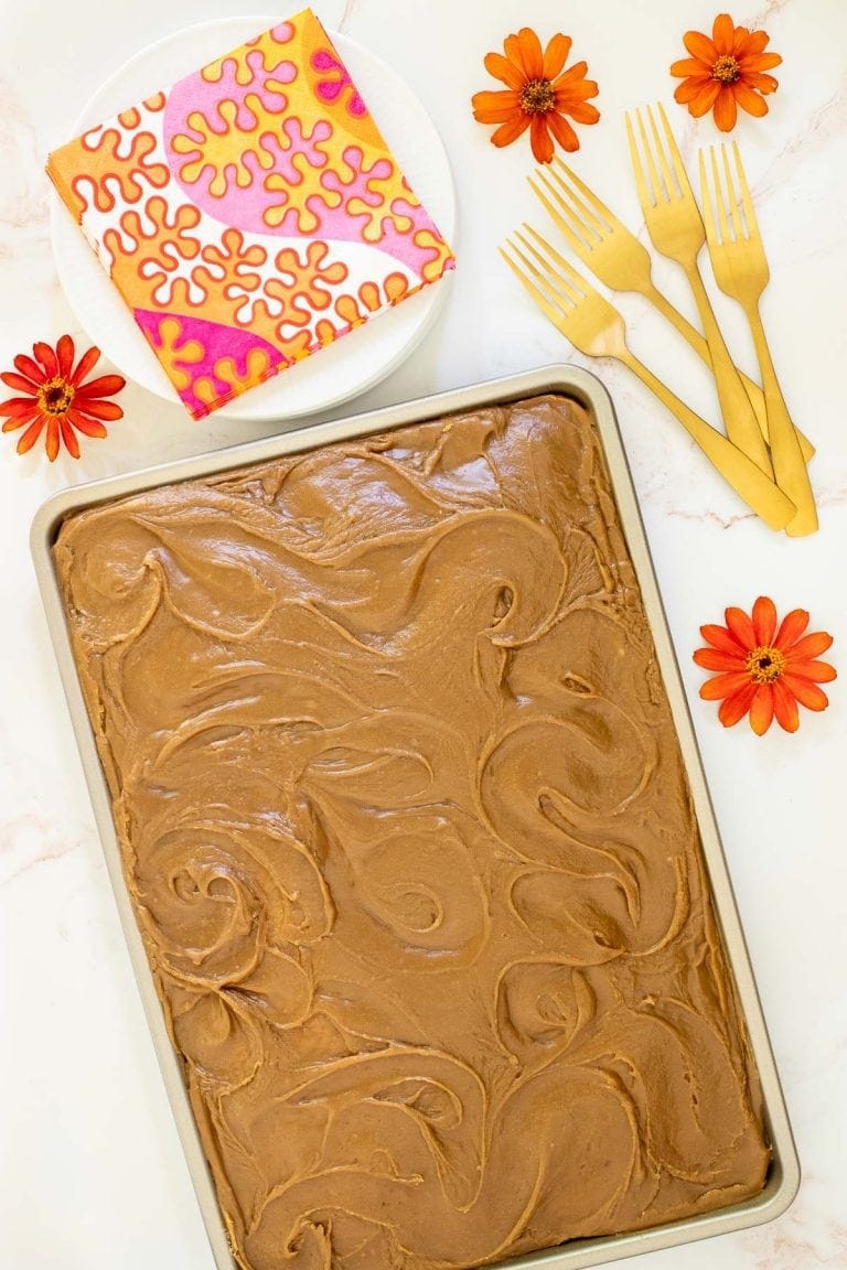 Vertical photo of a Ridiculously Easy Caramel Buttermilk Sheet Cake on a white marble surface. The photo has an orange border and the cake is surrounded by orange zinnias.