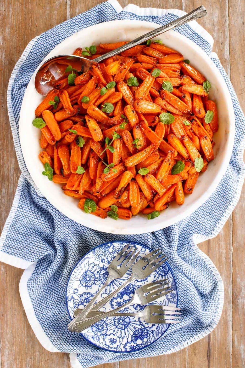 Vertical overhead photo of Honey Ginger Charred Carrots in a white serving bowl surrounded by a blue and white patterned towel.
