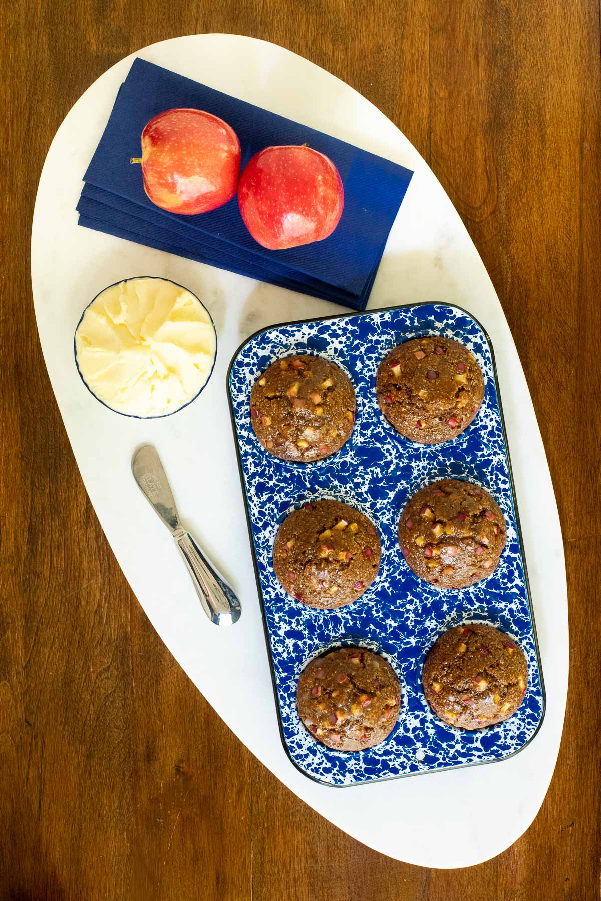 Vertical overhead photo of Honey-Glazed Apple Bran Muffins in a blue and white baking tin with apples and butter on the side.